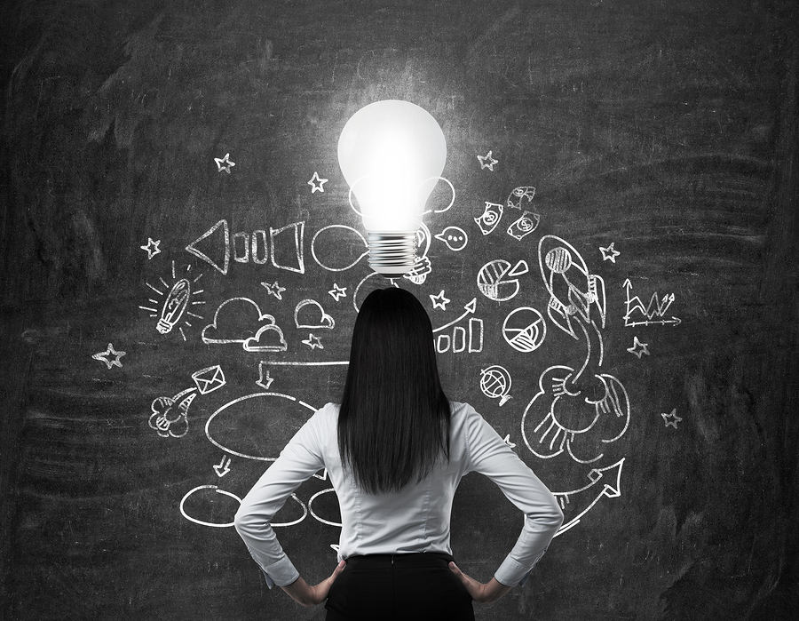 Rear view of the brunette business lady who is looking for new business ideas. Drawn business icons on the black chalk board and a light bulb as a concept of new idea.